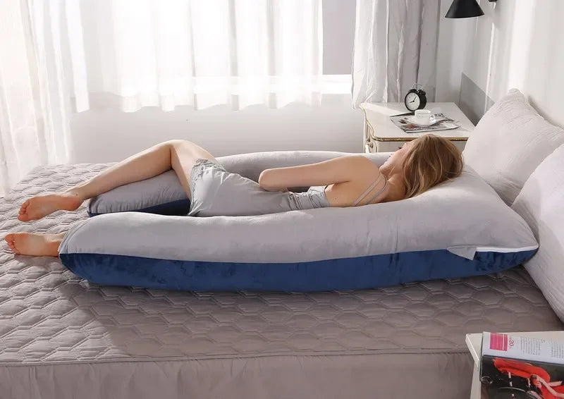 Pregnant woman lying on a large grey and blue U-shaped body pillow in a bedroom.