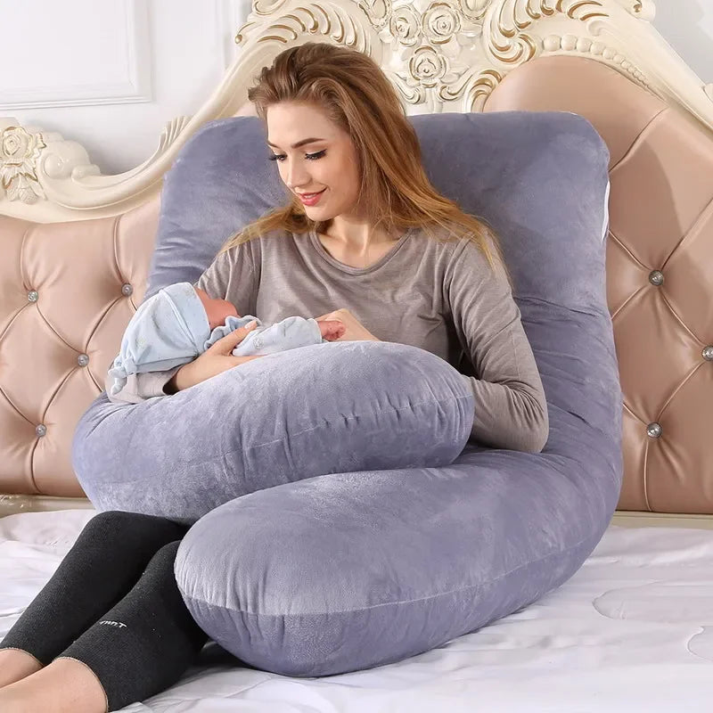 Mother holding a baby while sitting on a U-shaped gray pillow in a bedroom.