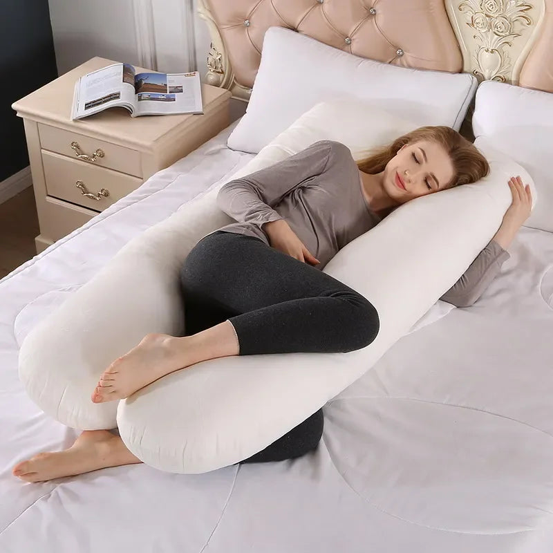 Pregnant maternity woman lying sideways on a white U-shaped body pillow in a bedroom.