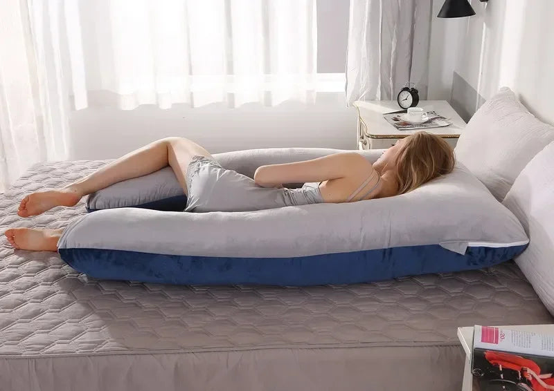 Pregnant maternity woman lying sideways on a grey and blue U-shaped body pillow in a bedroom.