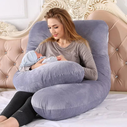 Mother holding a baby while sitting on a U-shaped gray pillow in a bedroom.